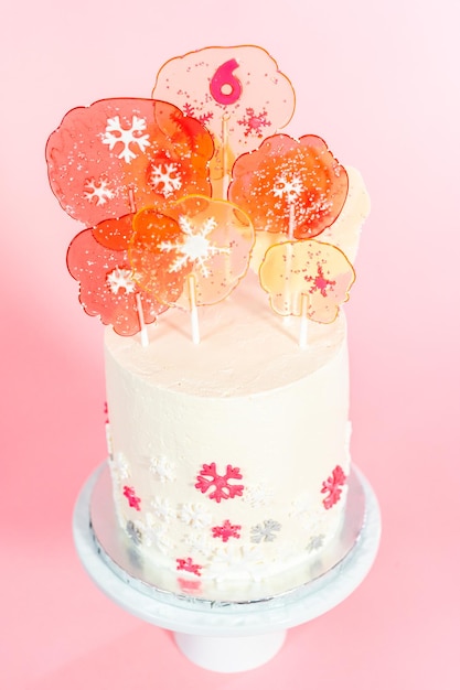 Torta rotonda alta con glassa di crema al burro italiana decorata con fiocchi di neve fondente e sormontata da grandi lecca-lecca rosa e bianchi su sfondo rosa.