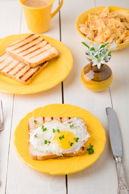 Tosti con l'uovo in piatto giallo vicino al vaso con il fiore su fondo di legno bianco.