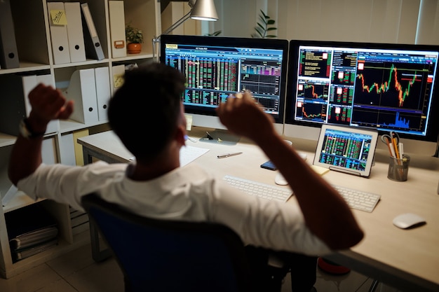 Trader che si allunga e si rilassa sulla sedia a tarda notte dopo aver terminato la sessione di trading, vista da dietro