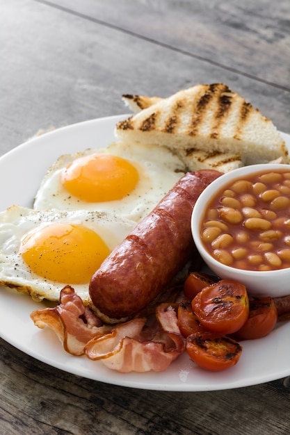 Tradizionale colazione inglese completa con uova fritte, salsicce, fagioli, funghi, pomodori grigliati e pancetta su superficie di legno