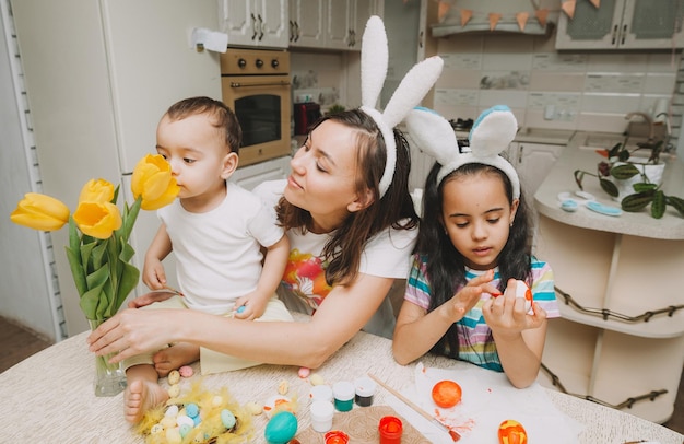 Tradizioni familiari di Pasqua Giovane madre amorevole che insegna ai bambini come dipingere le uova per Pasqua seduti insieme al tavolo della cucina