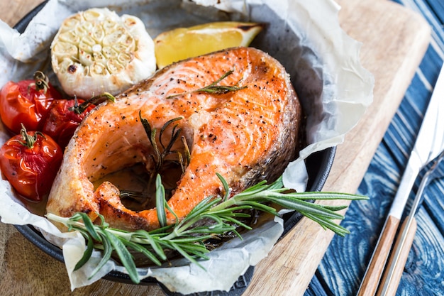 Trancio di salmone al forno con rosmarino, limone e spezie in padella nera su fondo rustico blu. Dieta bilanciata