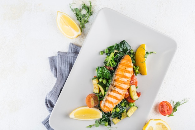 Trancio di salmone con verdure fresche e fette di limone su un piatto bianco con un tovagliolo su sfondo chiaro. Vista dall'alto con uno spazio di copia per il testo. Cibo da ristorante. Orientamento orizzontale.