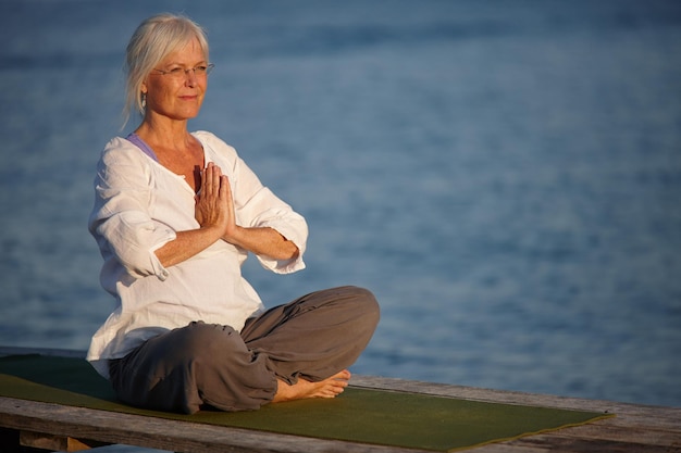 Tranquillità dall'oceano Scatto di una donna matura attraente che fa yoga su un molo sull'oceano