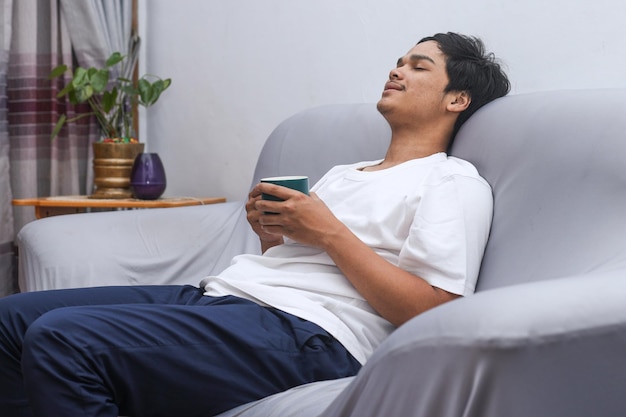 Tranquillo giovane asiatico seduto sul divano con gli occhi chiusi che beve caffè caldo a casa
