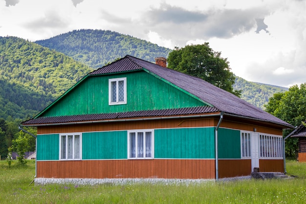 Tranquillo paesaggio rurale estivo in giornata di sole luminoso. Illuminata dal sole bella casa residenziale in legno dipinta nei colori verde, blu e marrone sul prato erboso in fiore sulle montagne boscose.