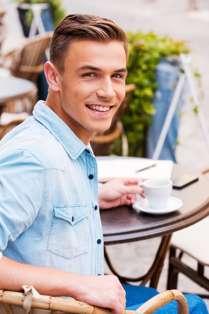 Trascorrere del buon tempo al caffè. Vista posteriore di un giovane allegro che beve caffè e guarda sopra la spalla mentre è seduto nel caffè sul marciapiede