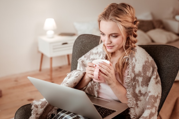 Trascorrere la giornata lavorativa. Signora calma e tranquilla con acconciatura complicata che abbraccia la tazza di caffè mentre guarda il video sul laptop