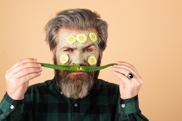 Trattamento facciale. Uomo barbuto divertente con maschera facciale di argilla in beauty spa. Ragazzo divertente con maschera facciale con cetriolo e aloe vera.