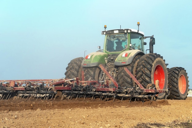 Trattore con un aratro che lavora nel campo Un agricoltore che guida un nuovo trattore moderno ara la terra La stagione della semina Primo piano rosso del trattore delle ruote enormi sullo sfondo di sabbia e polvere