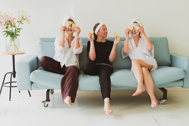 Tre amiche, giovani donne con maschere cosmetiche, si prendono cura del loro viso sul divano di casa e si divertono