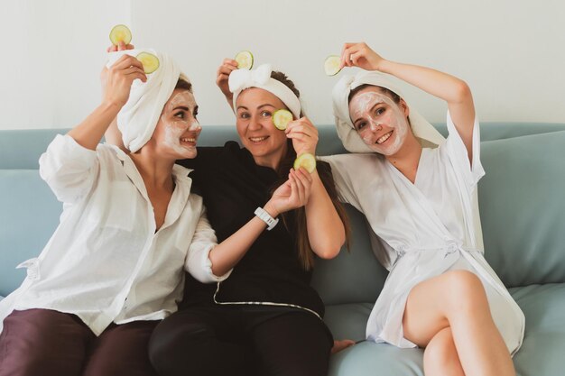 Tre amiche, giovani donne con maschere cosmetiche, si prendono cura del loro viso sul divano di casa e si divertono