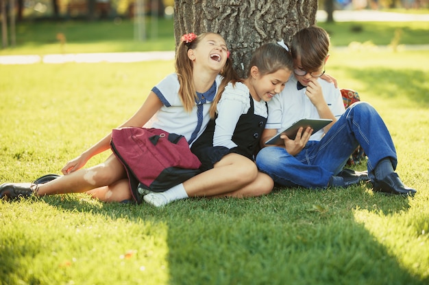 Tre amici di scuola adolescenti sorridenti che si siedono nel parco sull'erba e che giocano insieme il nuovo gioco della compressa. Emozioni diverse sui loro volti