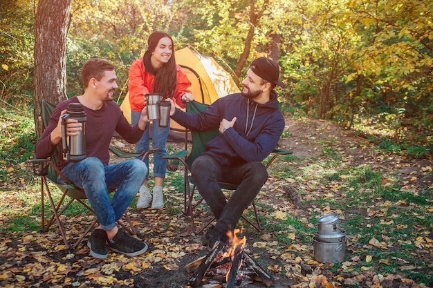 Tre amici sono insieme nella foresta. Tengono insieme le tazze e si guardano. Alcuni tengono il thermos nelle mani. Loro sono felici.