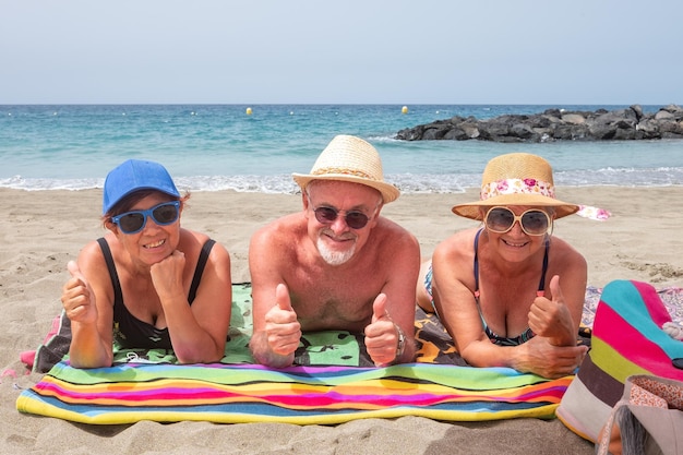 Tre anziani caucasici sorridenti con occhiali e cappelli sdraiati sulla spiaggia del mare a prendere il sole godendosi il tempo libero e le vacanze felici donne mature e uomo che guarda l'obbiettivo con thunbs up