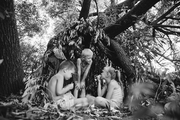 Tre bambini del villaggio giocano in una capanna che loro stessi hanno costruito con foglie e ramoscelli Casa di legno nella foresta Fotografia in bianco e nero