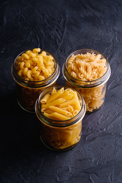 Tre barattoli di vetro con varietà di pasta cruda del grano dorato su fondo strutturato nero scuro, vista di angolo