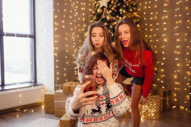 Tre belle ragazze fanno selfie al telefono, smorfie. Migliori amiche della giovane donna che celebrano il Natale in casa. Bellissime decorazioni natalizie dorate su un alto albero di Natale