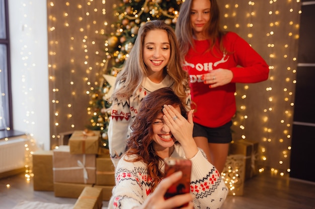 Tre belle ragazze fanno selfie al telefono, smorfie. Migliori amiche della giovane donna che celebrano il Natale in casa. Bellissime decorazioni natalizie dorate su un alto albero di Natale