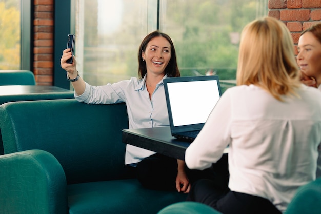 Tre bellissimi colleghi in una pausa caffè di lavoro. Ragazze attraenti con un laptop sono sedute a un tavolo, una di loro vuole fare un selfie. Concetto di affari, bellezza e istruzione.