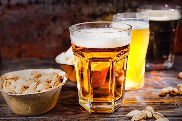 Tre bicchieri con birra chiara, non filtrata e scura stanno in fila vicino a piatti con snack e pistacchi sparsi sulla scrivania di legno scuro. Concetto di cibo e bevande