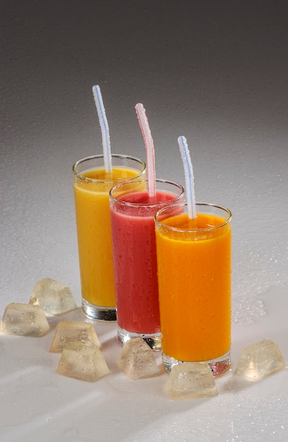 Tre bicchieri di succo di frutta con cannucce di plastica e ghiaccio su sfondo grigio sfumato.
