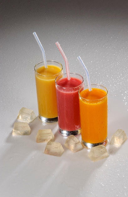 Tre bicchieri di succo di frutta con cannucce di plastica e ghiaccio su sfondo grigio sfumato.