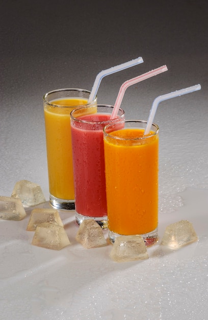 Tre bicchieri di succo di frutta con cannucce di plastica e ghiaccio su sfondo grigio sfumato.