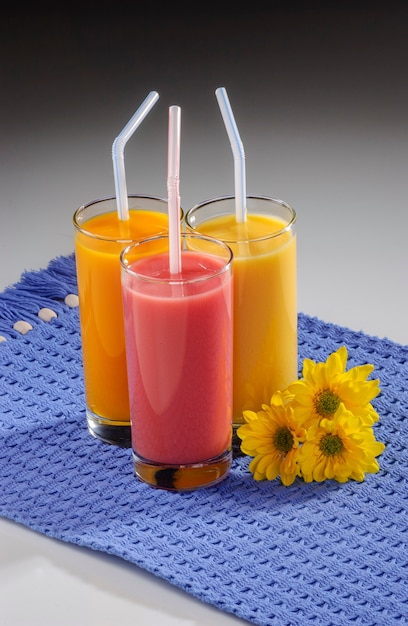 Tre bicchieri di succo di frutta con cannucce di plastica su tovaglietta blu su sfondo grigio sfumato.