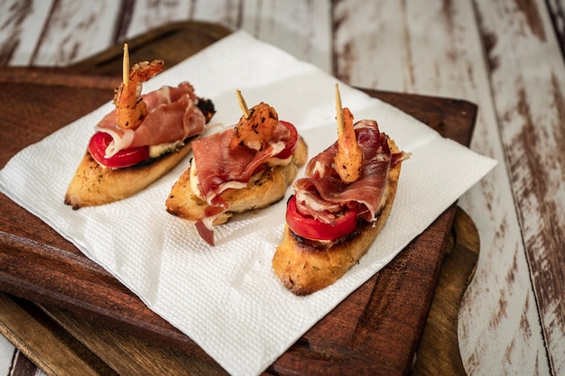 Tre bruschette con prosciutto serrano pomodoro ciliegino e gamberi alla griglia su una tavola di legno Concetto di cibo mediterraneo