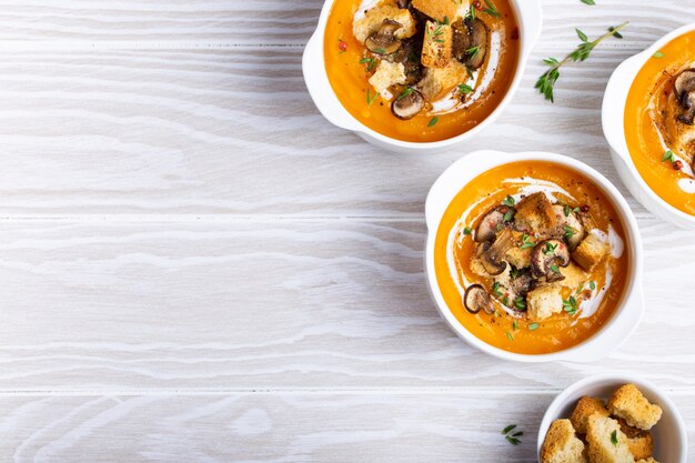 Tre ciotole in ceramica bianca con purea di zuppa di crema di zucca con funghi, crostini e panna su fondo rustico di legno bianco, vista dall'alto, primo piano. Cena in famiglia e comfort food, spazio per il testo