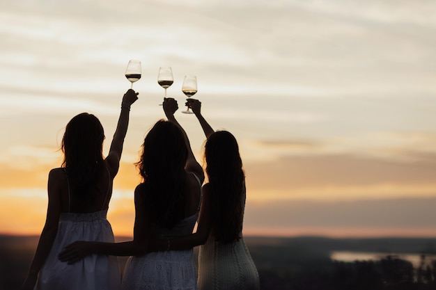 Tre donne vestite di bianco reggono bicchieri di vino e brindano con il sole che tramonta alle loro spalle.