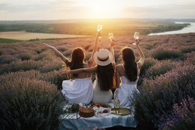 Tre donne vestite di bianco sono sedute su un tavolo da picnic in un campo di lavanda, con in mano bicchieri di vino e il sole sta tramontando all'orizzonte.