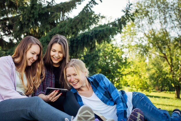 Tre giovani donne graziose sul prato nel parco guardano smartphone e ridono