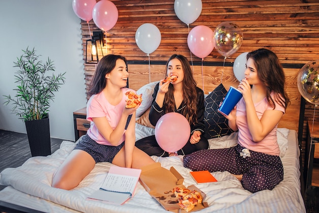 Tre giovani donne siedono sul letto nella sala. Mangiano pizza e bevono cola. le donne hanno una festa in pigiama. Loro sono felici.