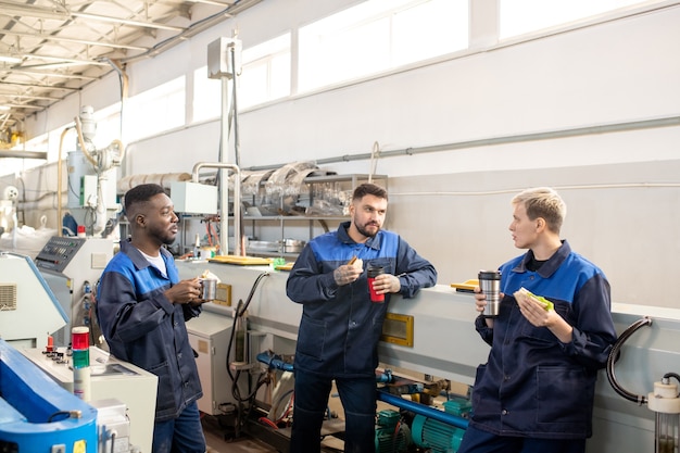 Tre giovani lavoratori interculturali in tuta in piedi contro la macchina industriale in una grande officina, discutendo i punti di lavoro e pranzando