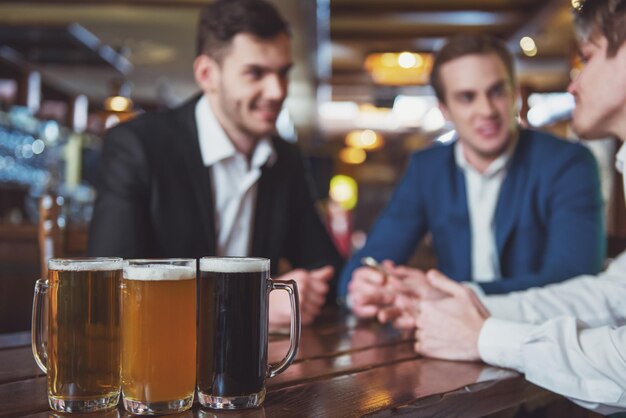 Tre giovani uomini d'affari in giacca e cravatta stanno sorridendo parlando e bevendo birra mentre si siedono in un pub si concentrano su bicchieri di birra