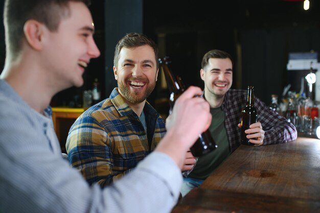 Tre giovani uomini in abiti casual sorridono tenendo bottiglie di birra mentre si trovano vicino al bancone del bar nel pub