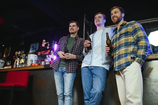 Tre giovani uomini in abiti casual sorridono tenendo bottiglie di birra mentre si trovano vicino al bancone del bar nel pub