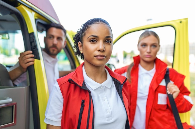 Tre paramedici multirazziali in piedi davanti al veicolo dell'ambulanza che trasportano attrezzature portatili