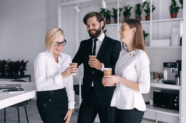 Tre partner allegri moderni ed eleganti durante una pausa che parlano durante il caffè in piedi sul posto di lavoro tenendo una tazza di caffè nelle loro mani uomo con setole