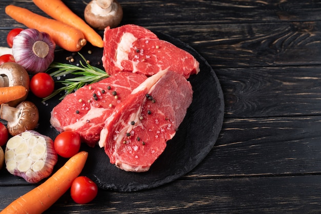 Tre pezzi di succosa carne cruda con verdure su un tagliere di pietra su un tavolo di legno nero