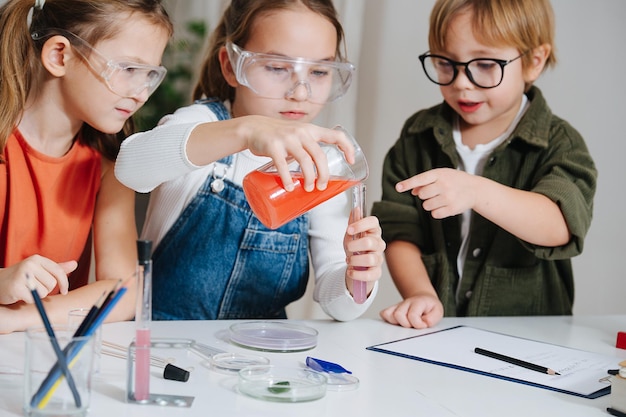 Tre ragazzini concentrati che svolgono un progetto di scienze domestiche, riempiendo con cura una fiaschetta dal bicchiere. Tutti dietro il tavolo, con gli occhiali. Vetreria chimica e liquidi colorati sul tavolo.