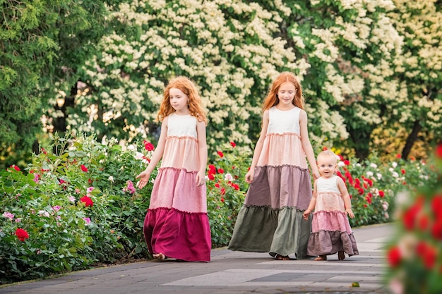 Tre sorelle dai capelli rossi in lunghi abiti di lino stanno passeggiando allegramente in giardino con le rose in una sera d'estate