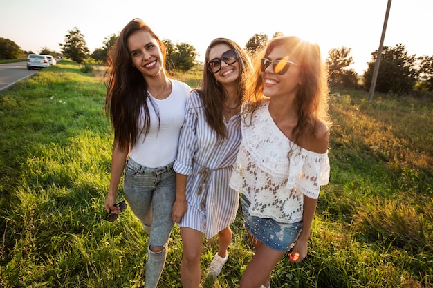 Tre splendide giovani donne in occhiali da sole vestite con bei vestiti stanno in campo e sorridono in una giornata di sole. .