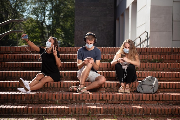 Tre studenti universitari seduti sulle scale che usano i loro smartphone indossando una maschera