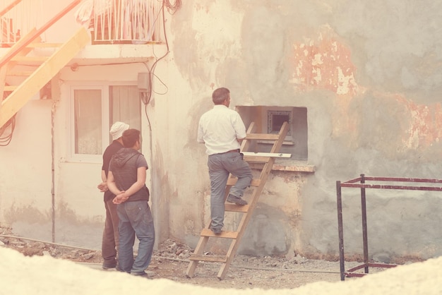 Tre uomini con un livello di costruzione e una scala in legno vicino al vecchio muro di pietra con una finestra tonica