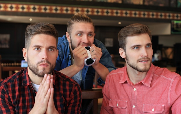 Tre uomini in abiti casual fanno il tifo per il calcio e tengono in mano bottiglie di birra seduti al bancone del bar in un pub.