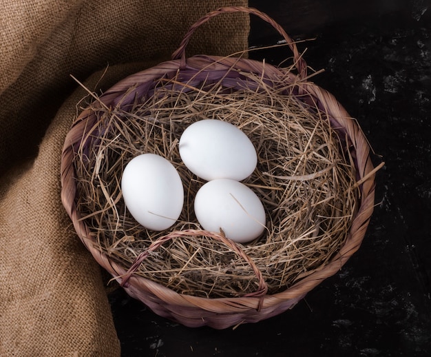 Tre uova di gallina in un cesto di fieno su un tavolo rustico.