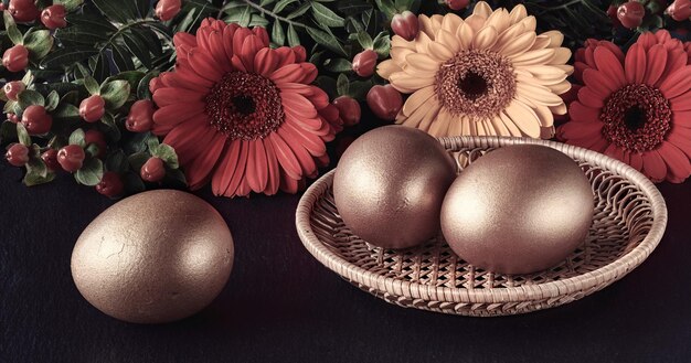 Tre uova di Pasqua dorate in piatto di rattan su sfondo scuro con fiori primaverili, margherita di erbe. Riflessi in vassoio di metallo.